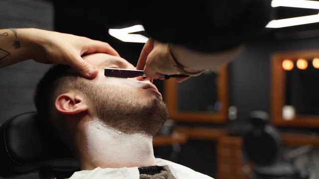 Professional barber shaves customer beard with straight razor. Beard cut with old-fashioned blade at barbershop. Handsome macho man getting his beard shaved in studio. Close-up shot.
