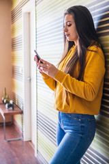 Young and attractive girl with long hair standing inside a house consulting the mobile phone or smartphone dressed in casual clothes