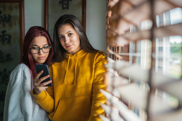 Two attractive young girls next to a window consulting the mobile phone or smartphone and laughing and talking to each other