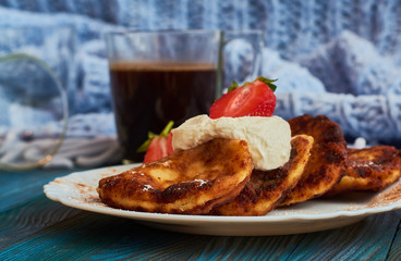 Delicious and useful breakfast of cheese pancakes with sour cream and strawberries cooked at home, with strong black coffee on blue background of wood and plaid.