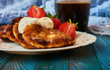 Delicious and useful breakfast of cheese pancakes with sour cream and strawberries cooked at home, with strong black coffee on blue background of wood and plaid.