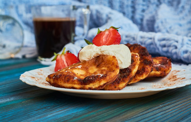 Delicious and useful breakfast of cheese pancakes with sour cream and strawberries cooked at home, with strong black coffee on blue background of wood and plaid.