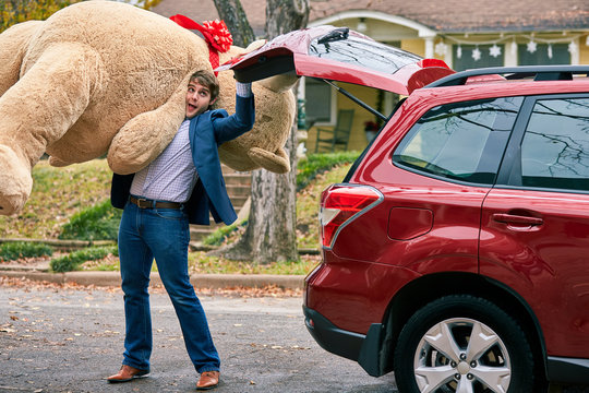 Man grabs a valentines day gift out of the trunk of his car, surprising his partner with a large teddy bear.