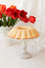 Festive homemade cakes. A round angel cupcake, a large biscuit or cake with a rose decoration on a white stand on a leg on a light background with a white vase and red tulips. Place for text.