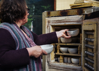 Frau legt mit der Hand Schalen zu vielen rustikalen Schalen aus Ton in einen Brennofen. Rohe...