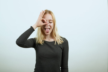 Young Blonde Woman In Black Sweater With Stylish Watch On White Background, A Silly Girl Shows Her Tongue And Made An Ok Sign With Fingers In Front Of Eye. The Concept Of Bold People