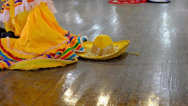 Traditional Mexican dance with woman wearing traditional Jalisco dress and man wearing mariachi costume. Yellow sombrero sits on the floor. View of dancers feet dancing on wooden stage. 