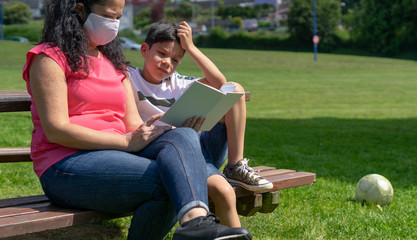 Mother reads her book to her son very concentrated in the park with a mask covering his mouth while he is bored and wants to play with the ball