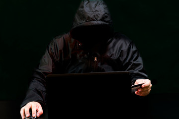 A hacker in a hood, without a face, on a black background sits at a laptop and enters data from a...