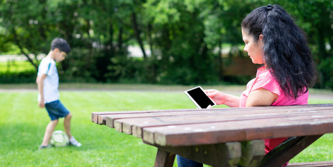 Fototapeta premium Mother reads her tablet very concentrated in the park while her child plays with the ball. Enjoy the outdoors.