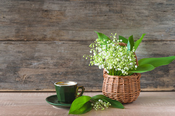 Greeting card. Bunch of lilies of the valley in wicker basket and vintage green cup of coffee on old wooden background. Space for text