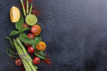 Fresh raw vegetables and herbs, asparagus, red cherry tomatoes, yellow tomatoes, lemon, lime, spinach, basil and spices on a black background. Top view, flatlay. Background image, copy space