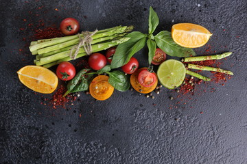 Fresh raw vegetables and herbs, asparagus, red cherry tomatoes, yellow tomatoes, lemon, lime, spinach, basil and spices on a black background. Top view, flatlay. Background image, copy space