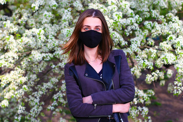 The girl in the protective medical mask is staying in front of a flowering white tree outdoors in the park