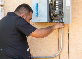 An electrical technician attaches metal conduit to a fuse box on the side of a house