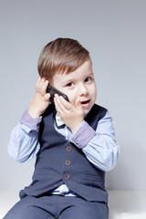 A boy in a shirt, bow tie, vest and trousers on a white background is talking on the phone. Stylish baby with hair styling