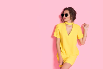 Beautiful girl wearing yellow dress and sunglasses posing on pink background in studio. Looking at camera.