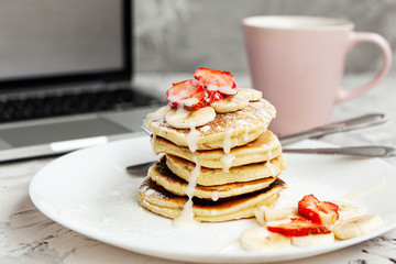 A stack of pancakes with fresh strawberries on a white plate on a light background. There is a laptop next to it. Work at home. Horizontal orientation. Homemade american pancakes. Copy space.