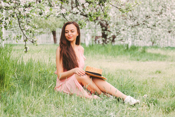 Portrait young carefree hipster girl in stylish pink dress sit on green grass on sunny countryside garden with blossom tree. Beautiful happy hippie woman on summer sunny meadow. Cheerful girl outdoors