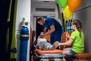 Unconscious mother lying on a stretcher in an ambulance car, a paramedic providing her first aid and talking to her little child © Anna Kosolapova