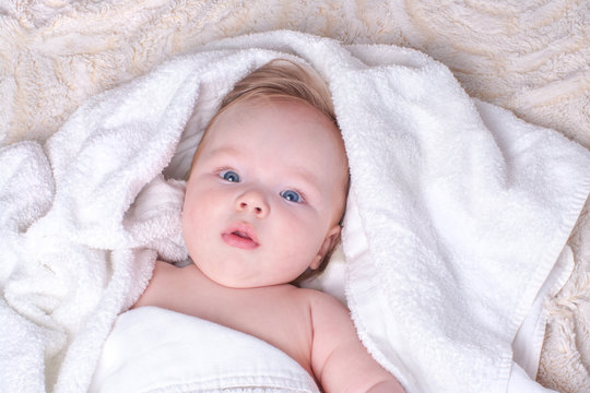 Surprised, newborn blue-eyed boy with plump lips and blond hair lies in dry, white towel on light fluffy blanket after bathing in bathroom. Textiles and bedding for children. Concept baby hygiene