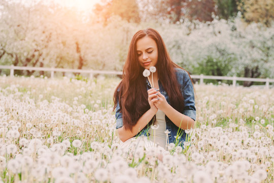 Portrait young beautiful hipster woman in denim jacket blow dandelions in hands while sit on green grass on garden. Happy carefree hippie girl relaxing on dandelion field. Spring flower pollen allergy