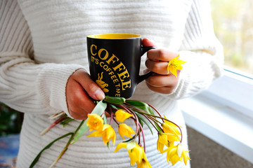 Hands holding a black and yellow mug with coffee and yellow meadow tulips on a window background. Home relaxation