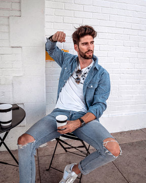 Handsome man in all jeans clothes, sitting alone in sidewalk cafe, holding a cup of coffee in his hand.
