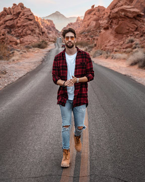 Man walking alone on the middle of the road, wearing plaid red shirt and sunglasses. Canyon road with red rocks are by the road.