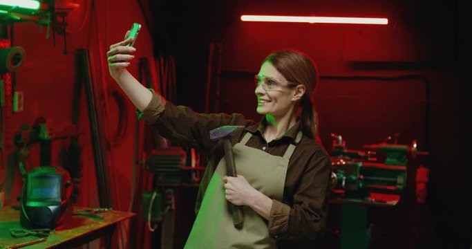 Caucasian beautiful woman in goggles and with hammer in hand posing to smartphone camera and making photo in metal workshop. Female steel worker taking selfie photos on mobile phone. Photography.