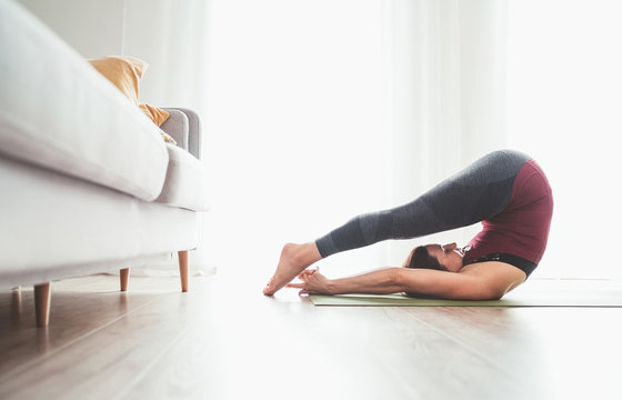 Young graceful Woman enjoying morning yoga exercises doing Halasana pose at home living room near the big window. Active people, body care and healthy lifestyle concept.