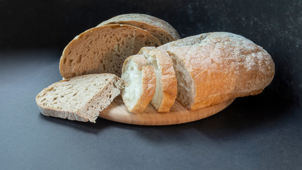Beautiful bread close-up, two types of bread made from wheat and rye flour on a round wooden board on a black background. Sliced and half a loaf.