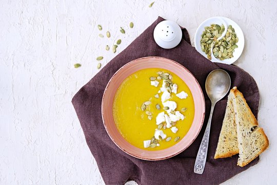 Pumpkin puree soup in a pink clay bowl on a light concrete background. Served with feta cheese, pumpkin seeds and croutons of white bread. Thanksgiving concept.