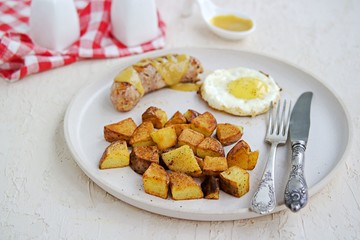 Large breakfast, home fries potatoes, pork sausage and fried egg on a white clay plate on a light concrete background. American food