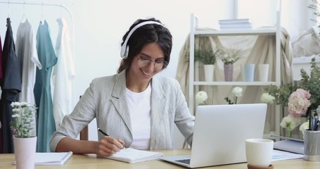 Smiling female wedding planner, fashion designer wearing headphones videoconferencing remote client on laptop, elearning watching training course, learning design style web class online, making notes. - Powered by Adobe