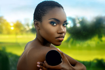 A sexy young black female with short black hair & beautiful makeup posing by herself in tropical surroundings while holding a box of powder foundation.