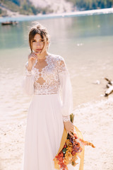 Fototapeta na wymiar Beautiful bride in a white dress with sleeves and lace, with a yellow autumn bouquet of dried flowers and peony roses, on the Lago di Braies in Italy. Destination wedding in Europe, on Braies lake.