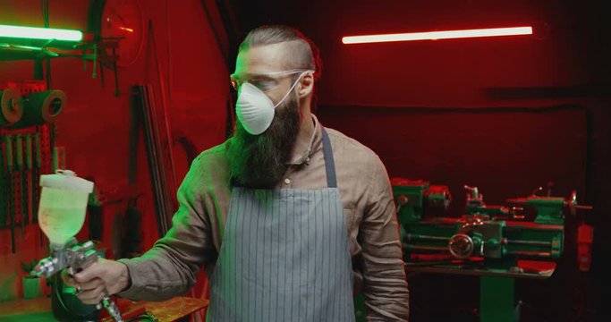 Man with beard in goggles, respiratory mask and apron working with pulverizator in workshop. Male metal worker painting steel with atomizer in hand. Paint spraying. Steel pulverization.
