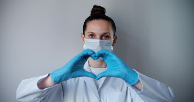 Woman with a medical mask and hands in latex glove shows the symbol of the heart. Doctor for the heart. Love to our pancreas. Love our medical professionals.