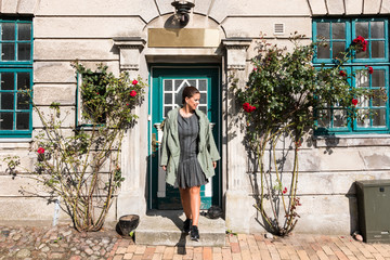 A woman leaves the house smiling with a green coat between two rose bushes. Lifestyle concept
