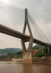Fototapeta premium Huangqikou, Chongqing, China - May 8, 2010: Yangtze River. 1 of 2 diamond shaped pylons of G319 suspension bridge with cables. Brown water and green hills with sprinkled white buildings in back.