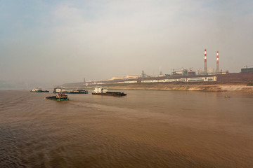 Fototapeta na wymiar Chongqing, China - May 8, 2010: Yangtze River. Giant power plant and factories on shoreline behind wide brown water with several coal transporting boats under light blue sky.