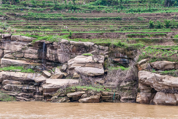 Fengdu, Chongqing, China - May 8, 2010: Yangtze River. Water streams over brown-gray rocks on shoreline, off terraced agriculture green plots behing brown water.