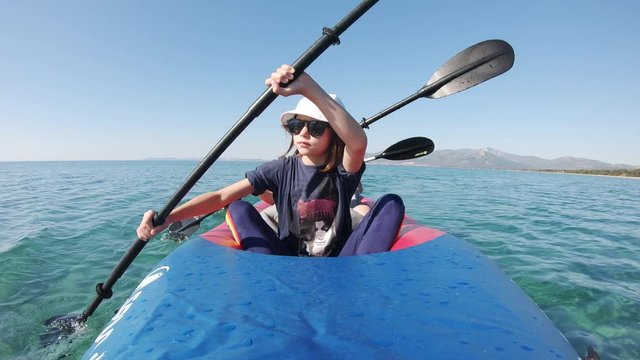 A young kid paddling a kayak with clear blue skies and mountains behind them.