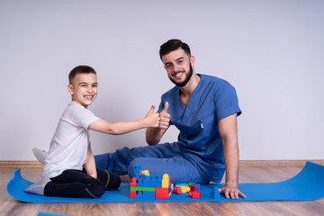 Young male doctor with beard in a blue uniform sitting on the floor next to the boy 10 years, they play educational toys, is looking at the camera and smile
