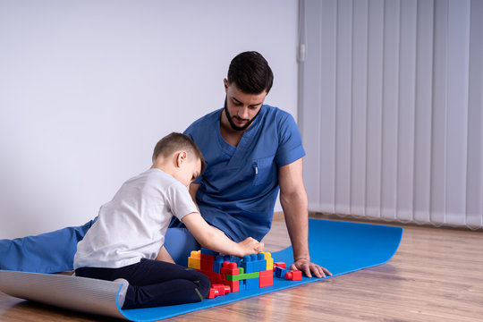 Young male doctor with beard in a blue uniform sitting on the floor next to the boy 10 years, they play educational toys, concept rehabilitation