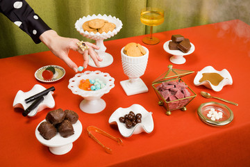 Still Life Spread of Cannabis Edibles in Milk Glass on Red Tablecloth with Hand Horizontal