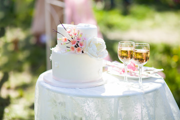 white wedding cake with flowers on a round table and two glasses of champagne, ceremony outside