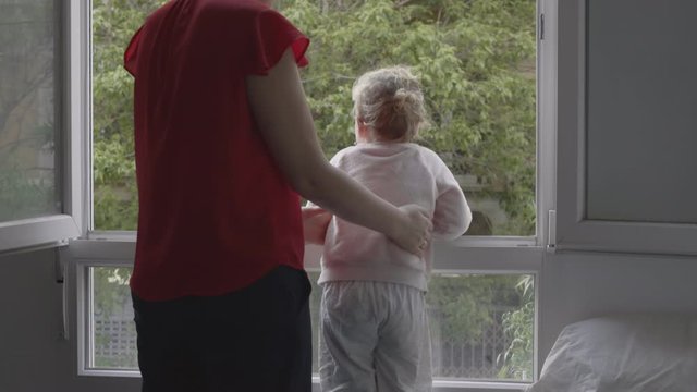 Mother Wants Little Daughter To Look Outside From The Window And Take The Air During Covid-19 Lockdown. Toddler Girl Having Fun And Dancing Making Mum Laugh