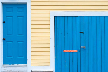 A bright blue house door and a garage door. On the garage door is a red sign with the words private property, lock, and latch. The doors are in a yellow wooden exterior wall of a building. 